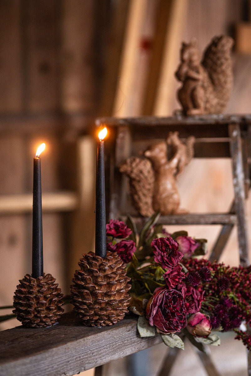 Två svarta ljusstakar formade som tallkottar med tända ljus, dekorerade med vinröda blommor | A Lot Decoration | Lilla Hem