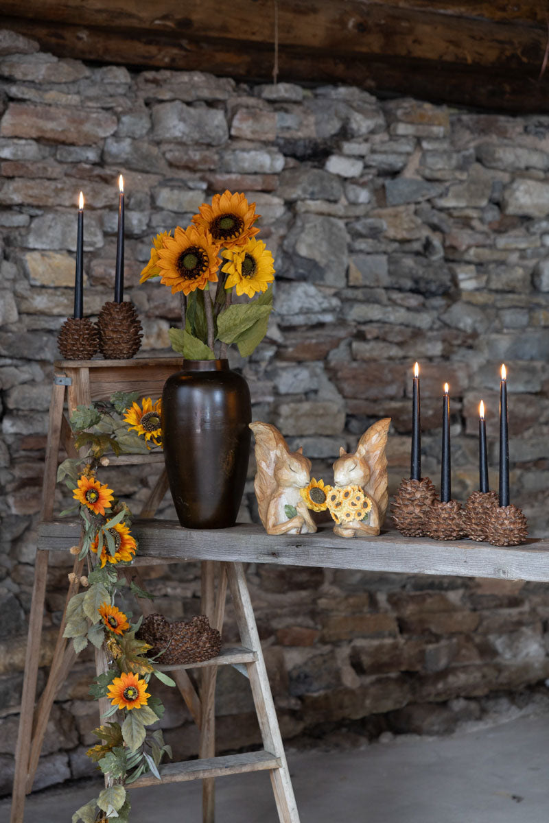 Ljusstake i form av tallkotte med brinnande svart ljus, ställd på hylla med solrosor och stenhöger | A Lot Decoration | Lilla Hem
