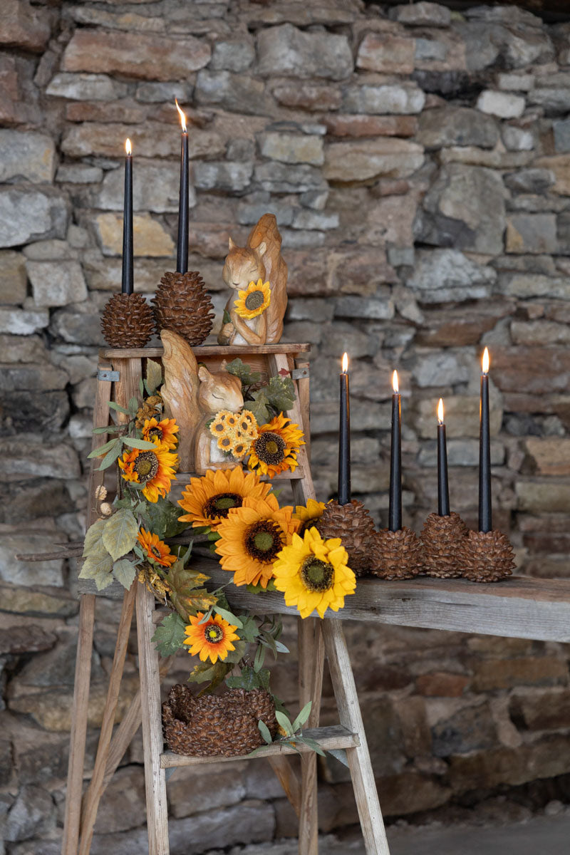 Ljusstake av tallkottar med svarta ljus, omgiven av solrosor och träfigurer mot stenmur | A Lot Decoration | Lilla Hem