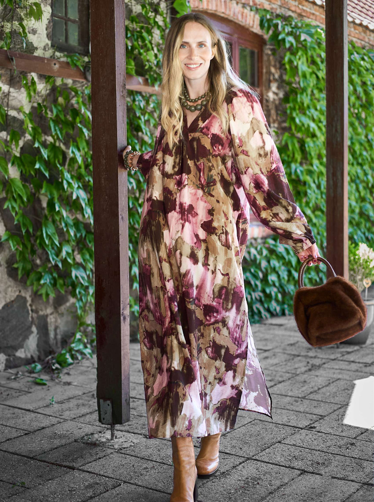 Modell bär blomstrig klänning i vinrött och rosa sett framifrån framför växtbeklädda tegelväggar | Black Colour | Lilla Hem
