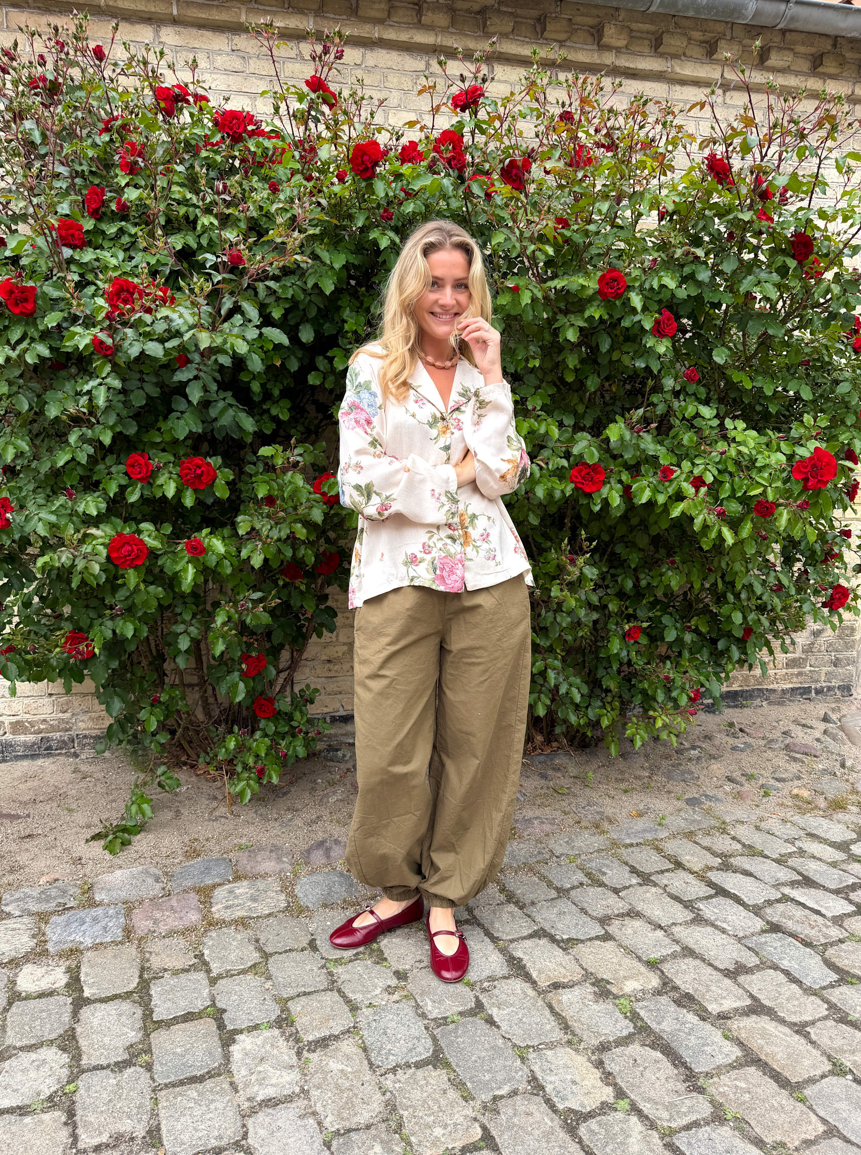 Modell bär blommig skjortblus i vitt framför röda rosor mot tegelvägg | Black Colour | Lilla Hem