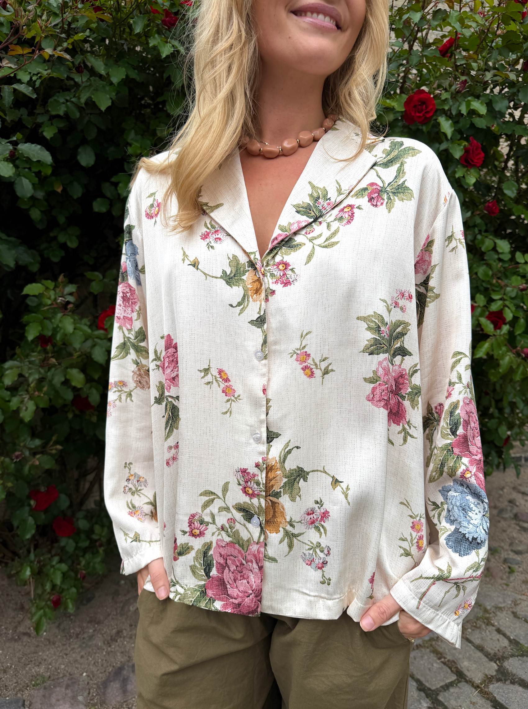 Modell bär skjorta med blommigt tryck framifrån mot rosenbusksbakgrund | Black Colour | Lilla Hem
