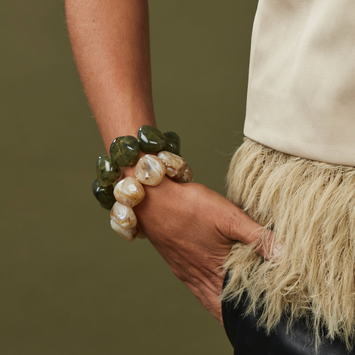 Närbild av armband med gröna och beige pärlor på handled, bärat med vit tröja och beige fransat plagg | Bud To Rose | Lilla Hem