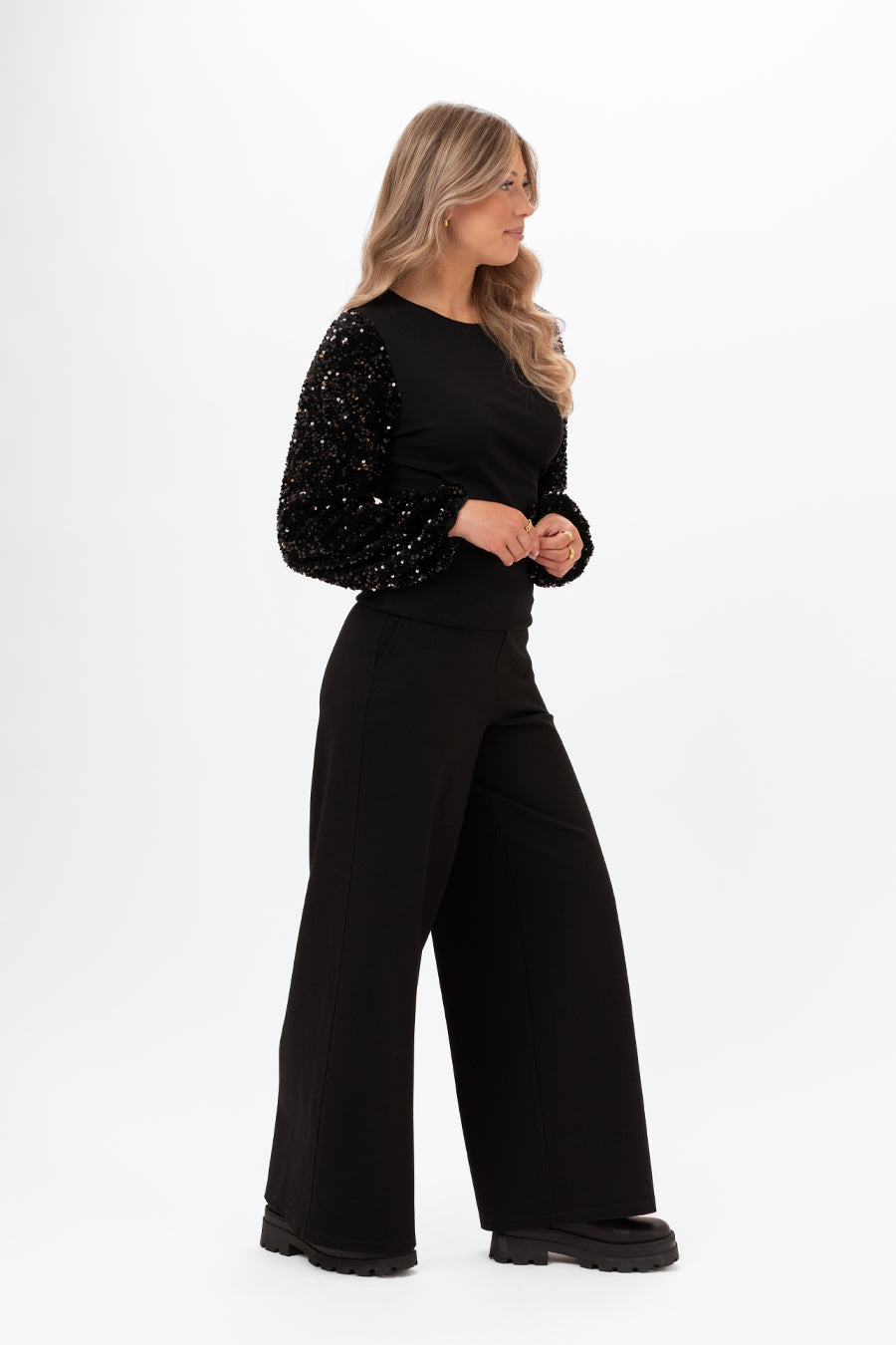 Modell bär svart topp med paljettärmarna sett från sidan, kombinerat med vida svarta byxor | Capri Collection | Lilla Hem