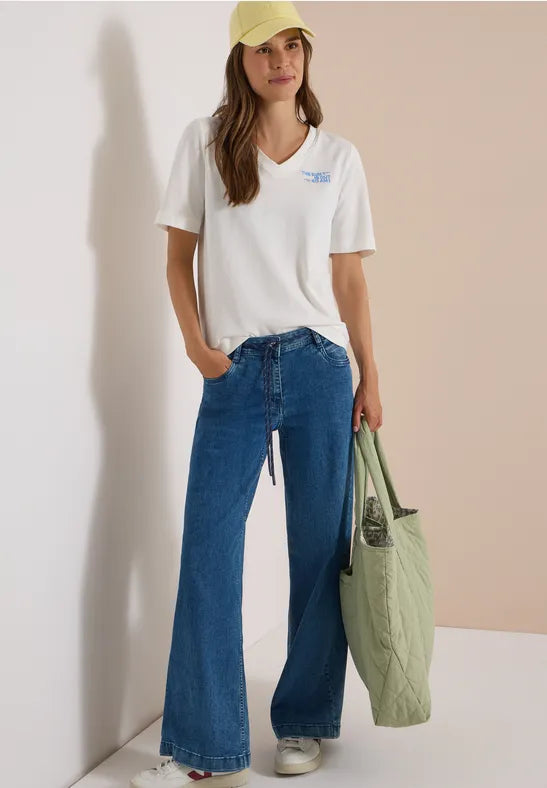 Modell bär vit t-shirt med blå tryck, vida blåa flare jeans och gul keps, sett framifrån | Cecil | Lilla Hem