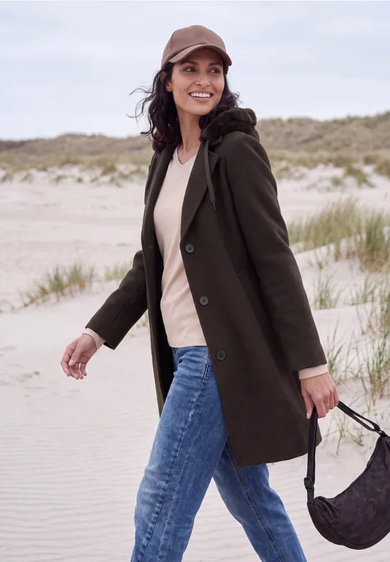 Modell bär mörkbrun kappa med päls över vit tröja och blå jeans, fotograferad i strandmiljö | Cecil | Lilla Hem