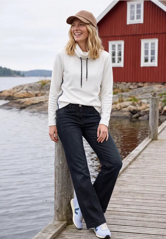 Modell bär vit hoodie och mörkblå jeans, fotograferad framifrån på brygga vid vatten | Cecil | Lilla Hem