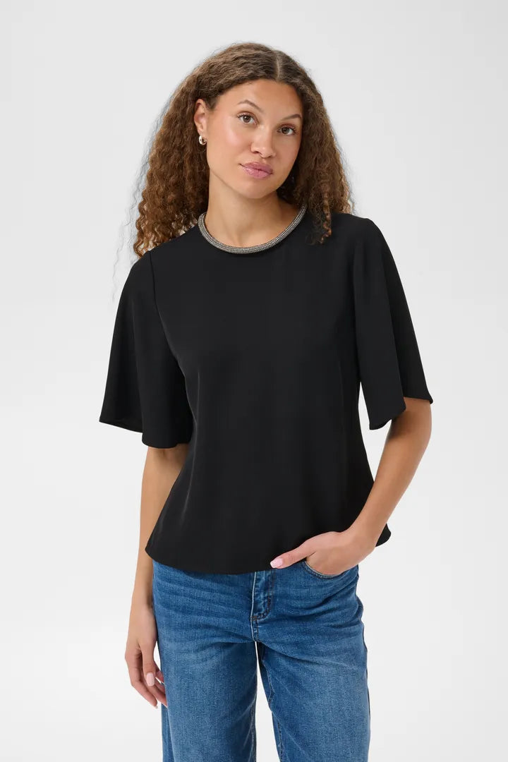 Modell bär svart blus med metallisk halsband, sett framifrån mot neutral bakgrund | KAFFE | Lilla Hem