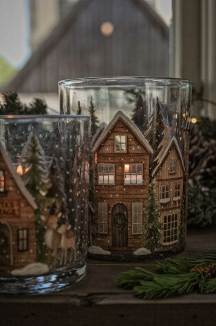 Två glaskrukor med tryckt vinterhusdesign och tänd belysning inuti, omgivna av granris | Majas Cottage | Lilla Hem