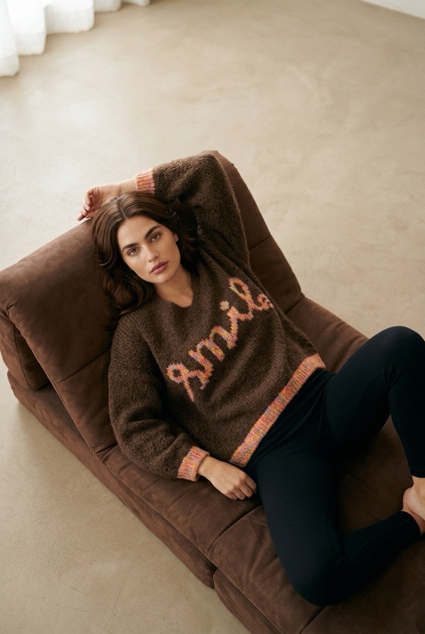 Modell sitter på brun stol och bär brun stickad tröja med rosa "SMILE"-text och ribbade kanter | L8 Fashion | Lilla Hem