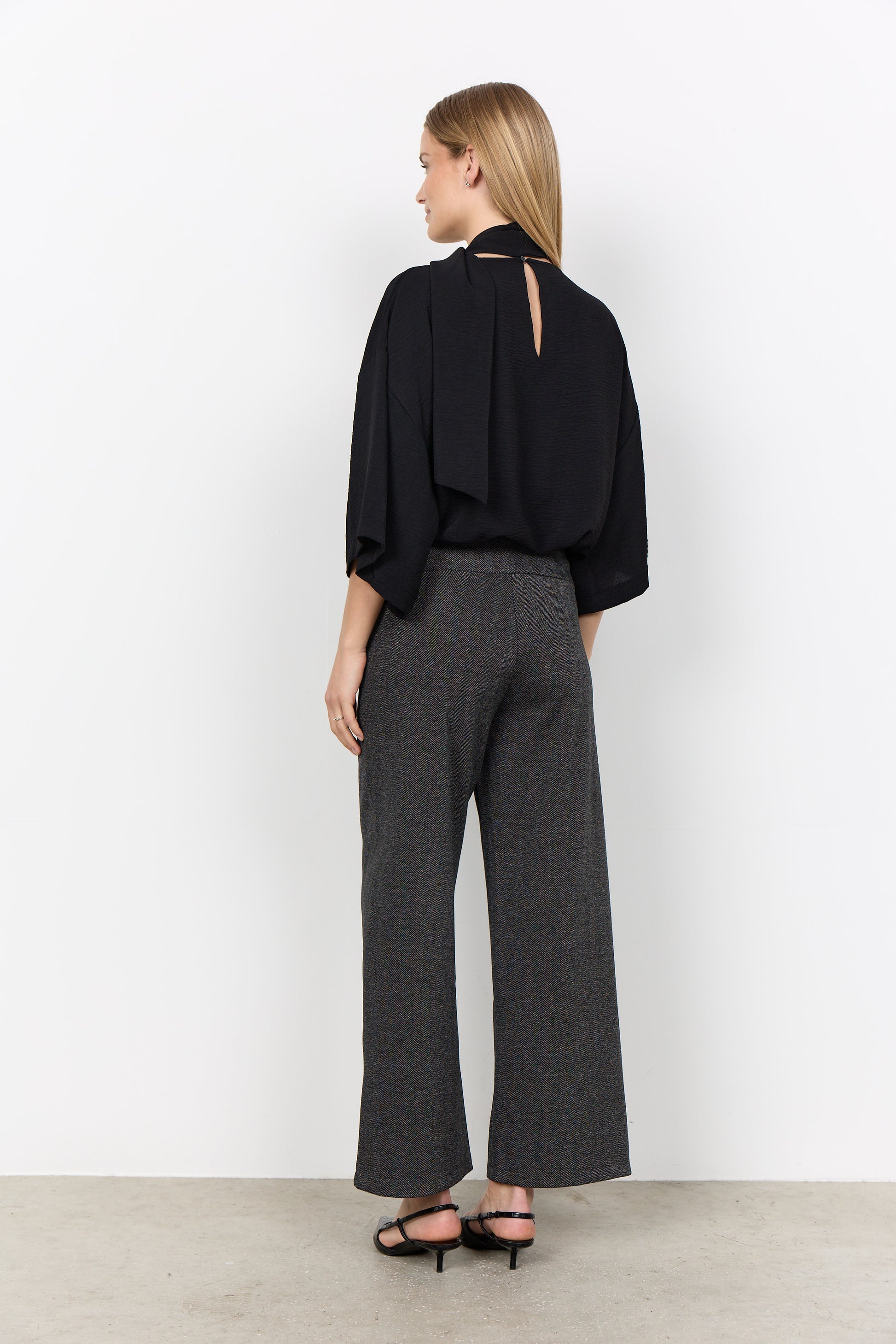 Modell bär svart blus med slits på ryggen och gråa vida byxor, sett från sidan bakifrån | Soya Concept | Lilla Hem