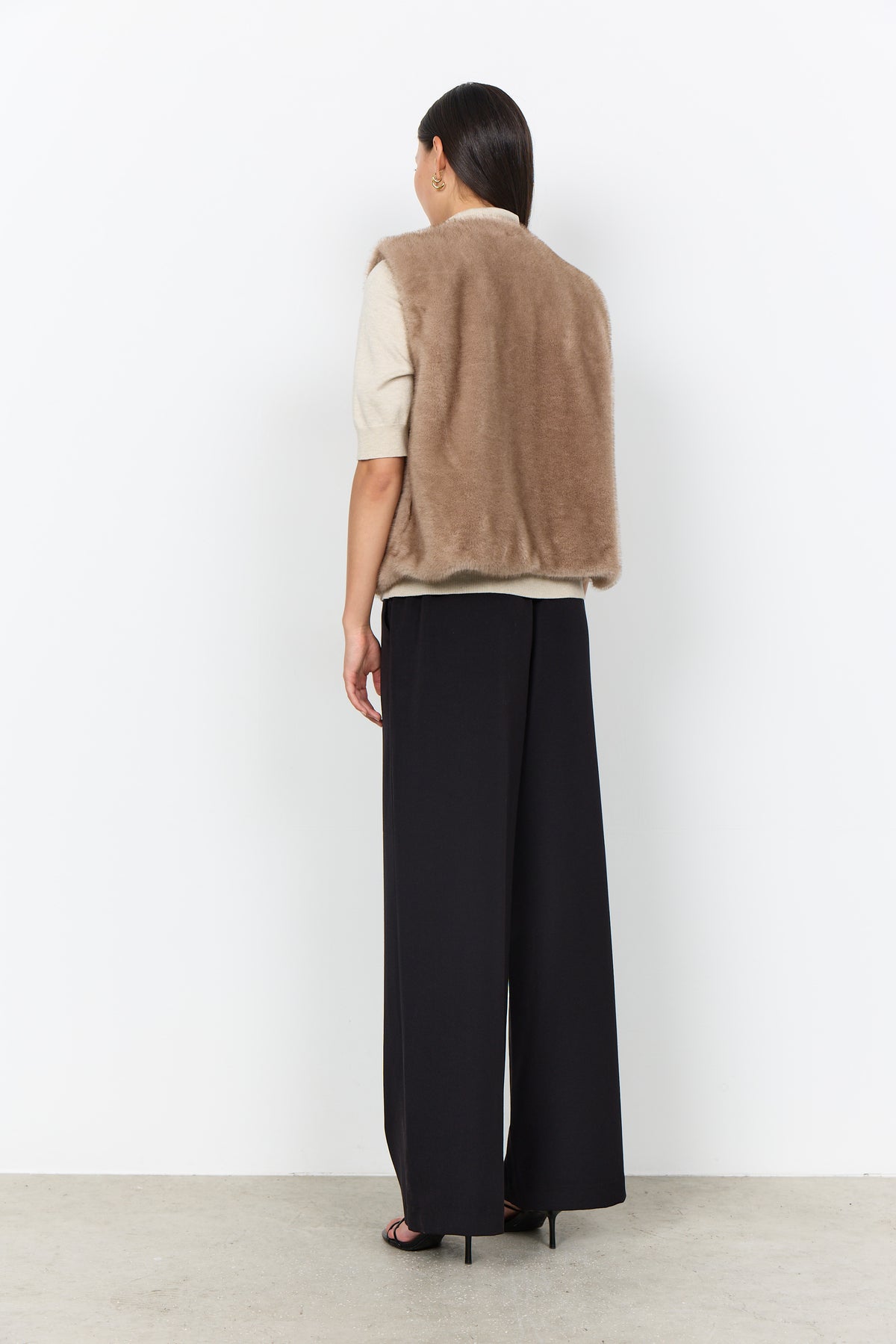 Modell bär beige pälsväst över vit tröja och svarta byxor, sett bakifrån | Soya Concept | Lilla Hem