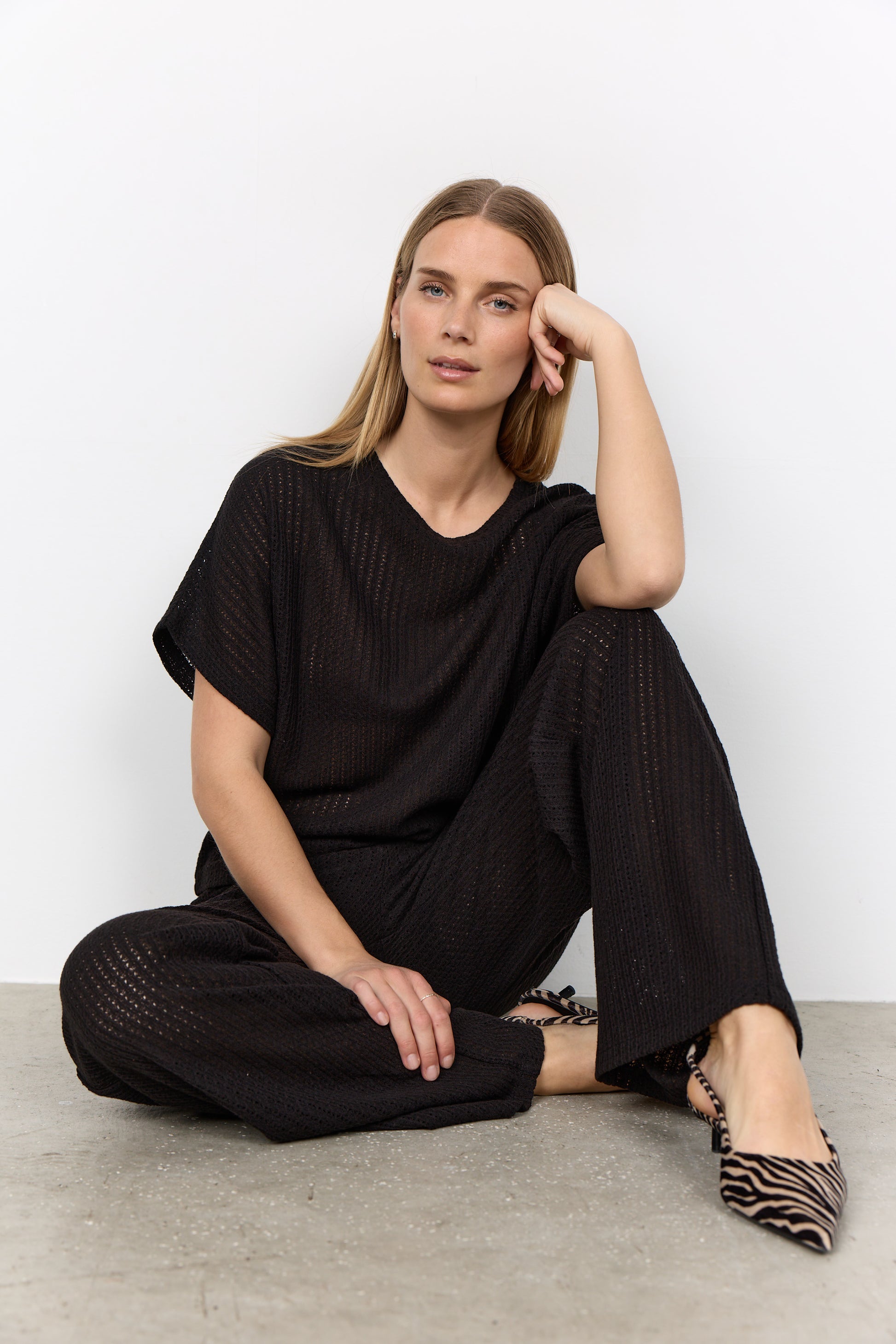 Modell sitter på golvet och bär svart ribbad t-shirt och byxor med v-ringning | Soya Concept | Lilla Hem