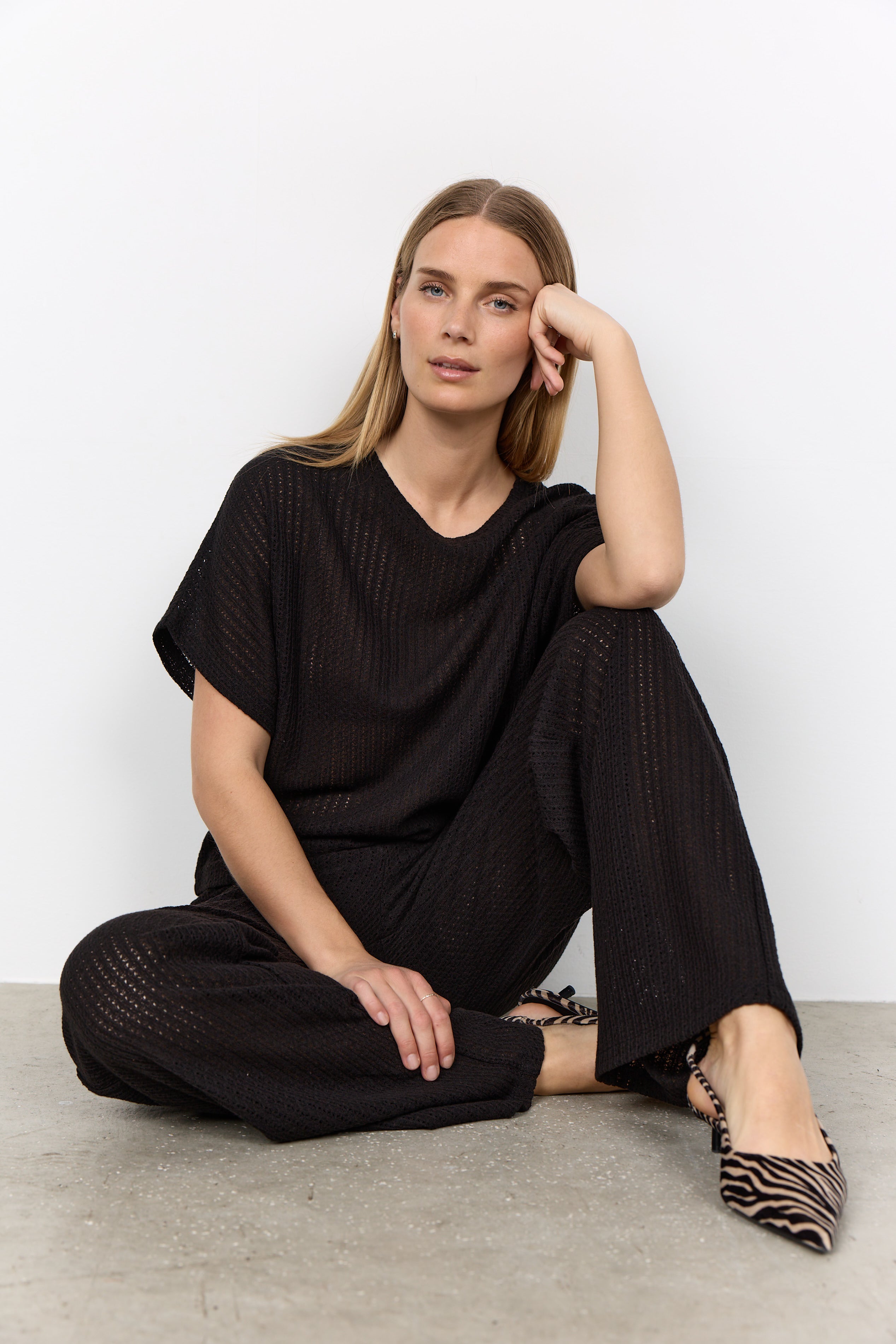 Modell sitter på golvet och bär svart ribbad t-shirt och byxor med v-ringning | Soya Concept | Lilla Hem